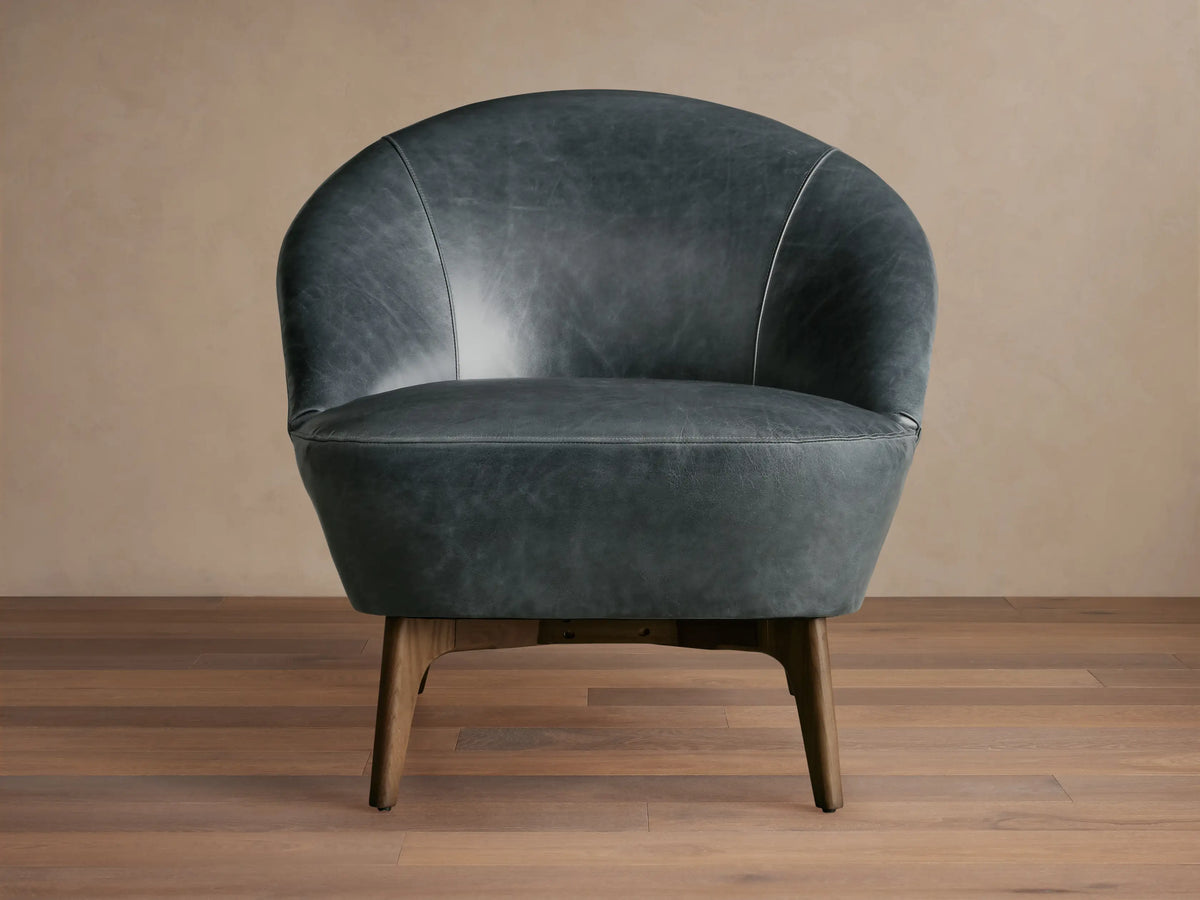 Elodie Leather Chair in dark grey leather with a rounded backrest and wooden legs, on a wooden floor with a beige wall backdrop.