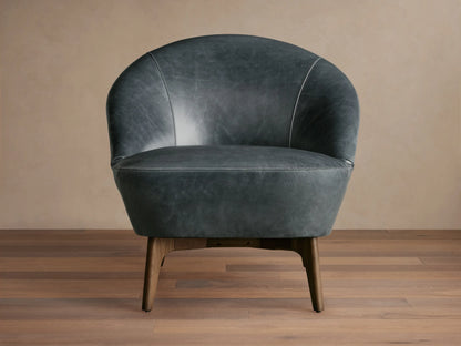 Elodie Leather Chair in dark grey leather with a rounded backrest and wooden legs, on a wooden floor with a beige wall backdrop.