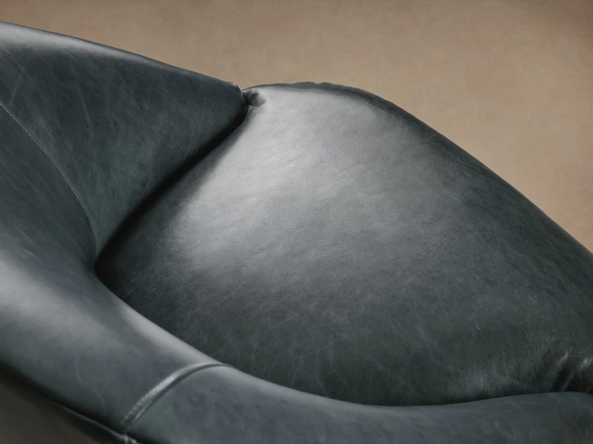 Elodie Leather Chair in dark gray leather with a rounded, cushioned backrest and seat, supported by four wooden legs on a wood floor.