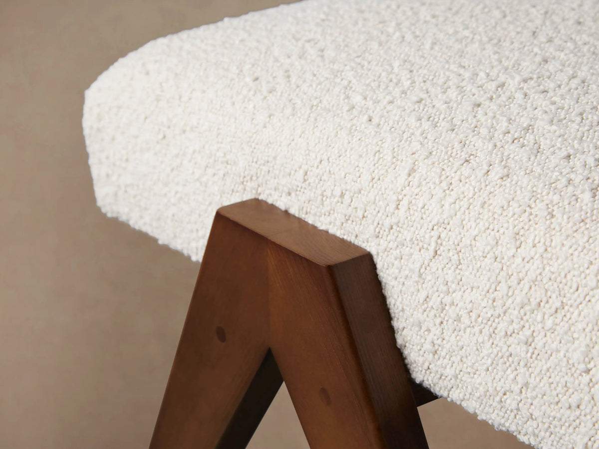 Elias Ottoman: Curved cream textured cushion, dark brown angled V-shaped wood legs, on a wood floor.