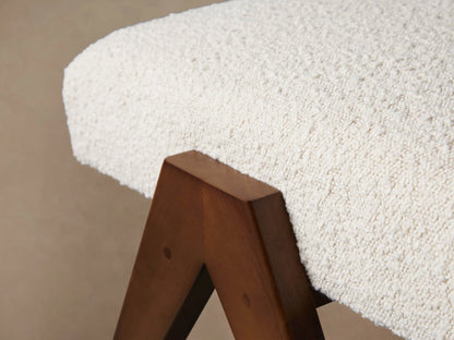 Elias Ottoman: Curved cream textured cushion, dark brown angled V-shaped wood legs, on a wood floor.