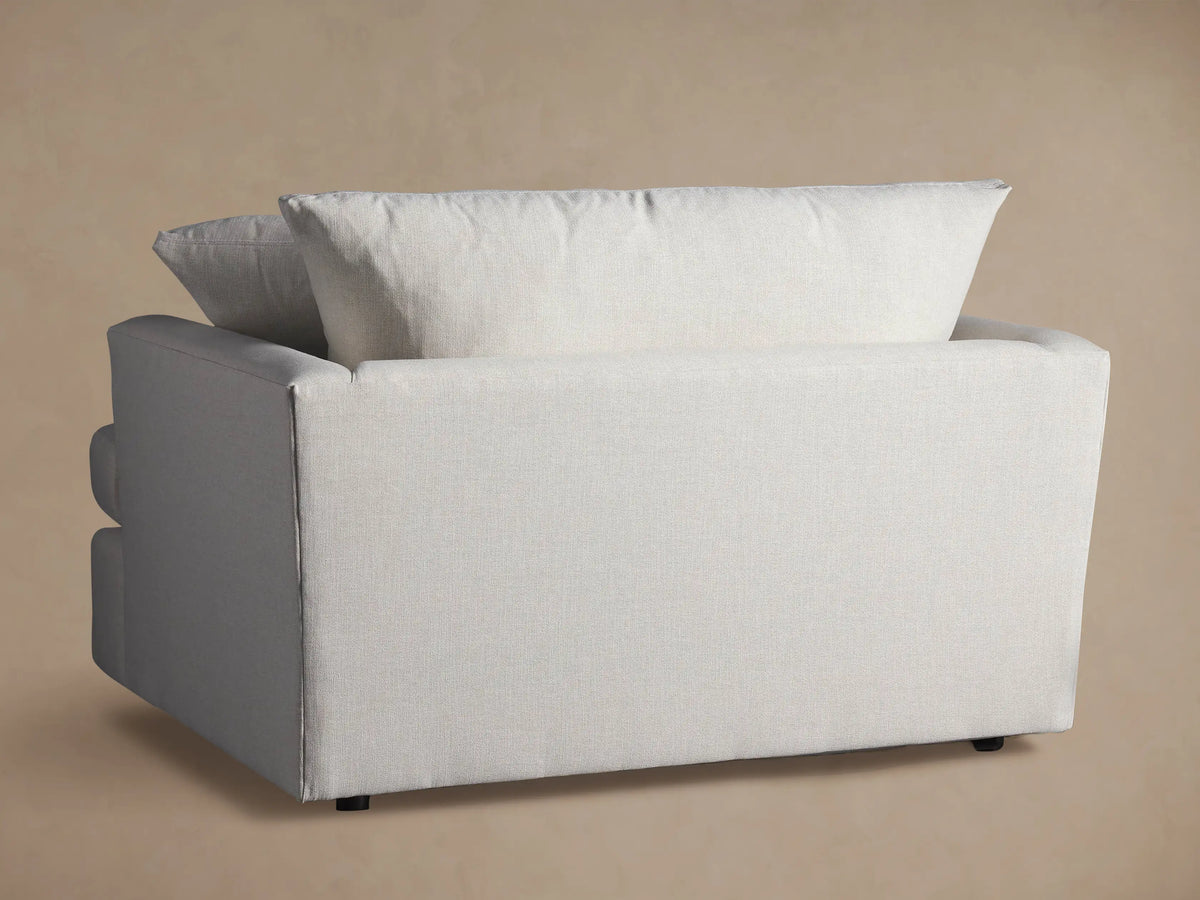 Close-up of Emory Chair's light, textured fabric armrest and cushions against a muted beige wall, highlighting its soft woven detail.