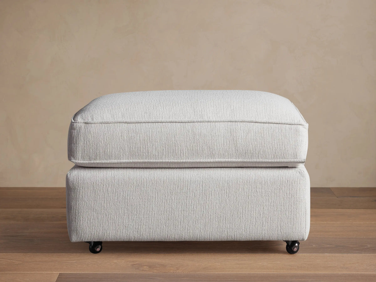Filmore Ottoman in light gray textured fabric, featuring a plush cushion top, on caster wheels on a wooden floor.