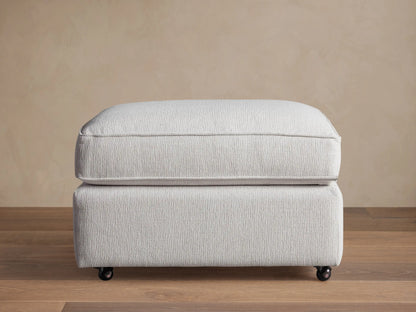 Filmore Ottoman in light gray textured fabric, featuring a plush cushion top, on caster wheels on a wooden floor.