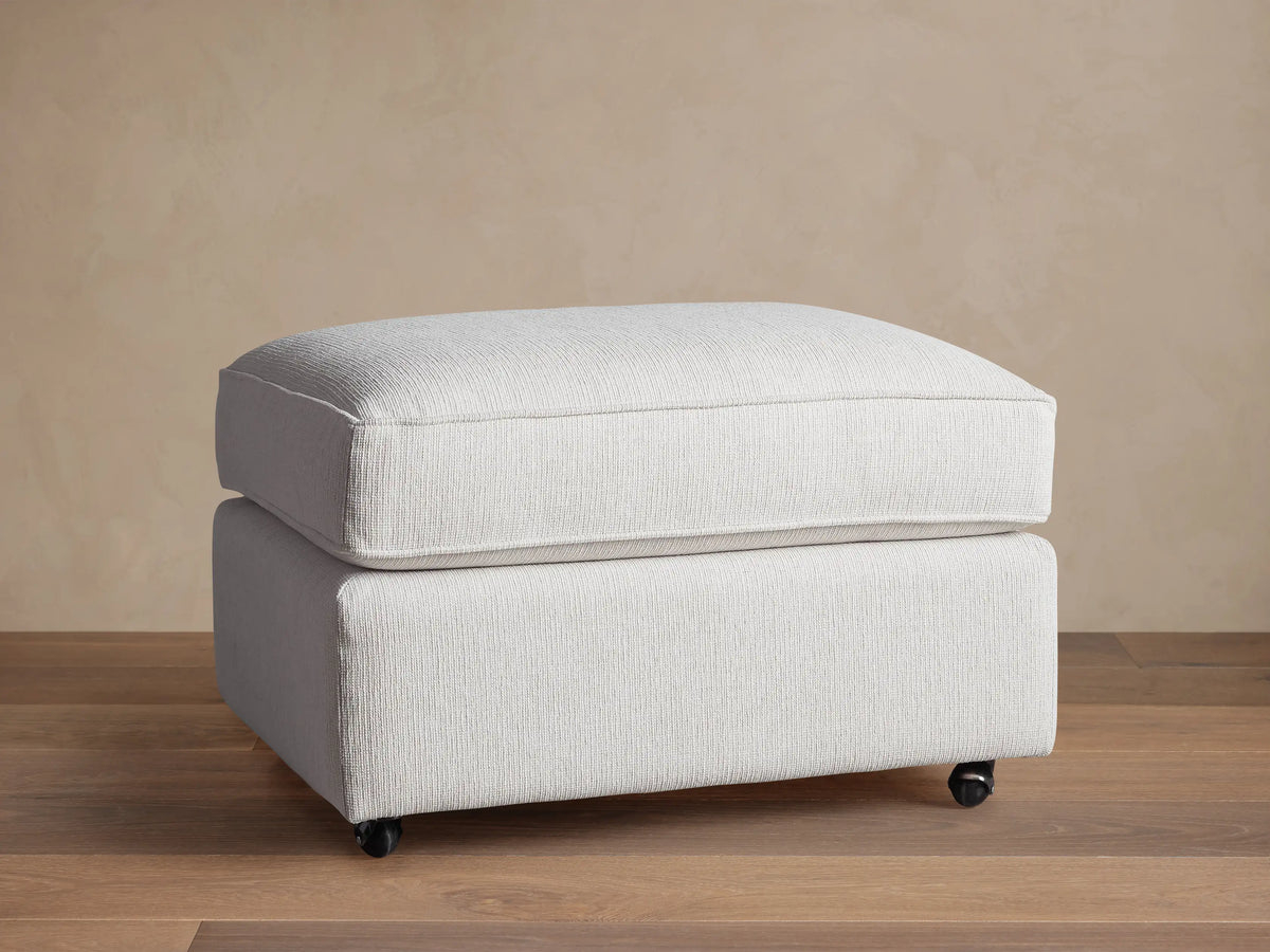 Filmore Ottoman in light gray textured fabric, featuring a plush cushion top, on caster wheels on a wooden floor.