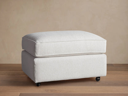 Filmore Ottoman in light gray textured fabric, featuring a plush cushion top, on caster wheels on a wooden floor.