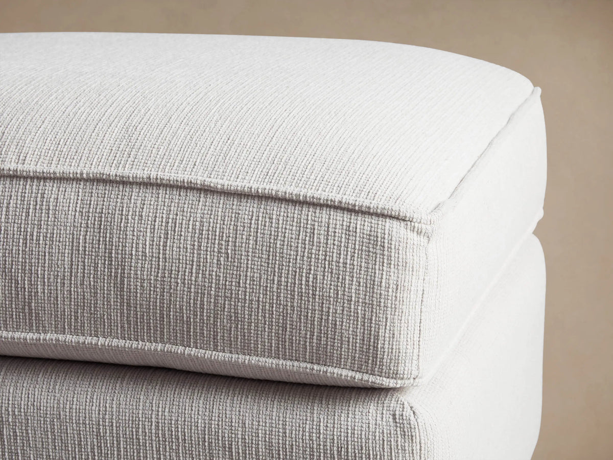 Filmore Ottoman, a light grey textured fabric rectangular ottoman with a plush cushion top, on a solid base with black castors.