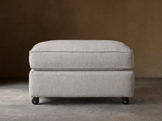Filmore Ottoman in light gray textured fabric, featuring a plush cushion top, on caster wheels on a wooden floor.