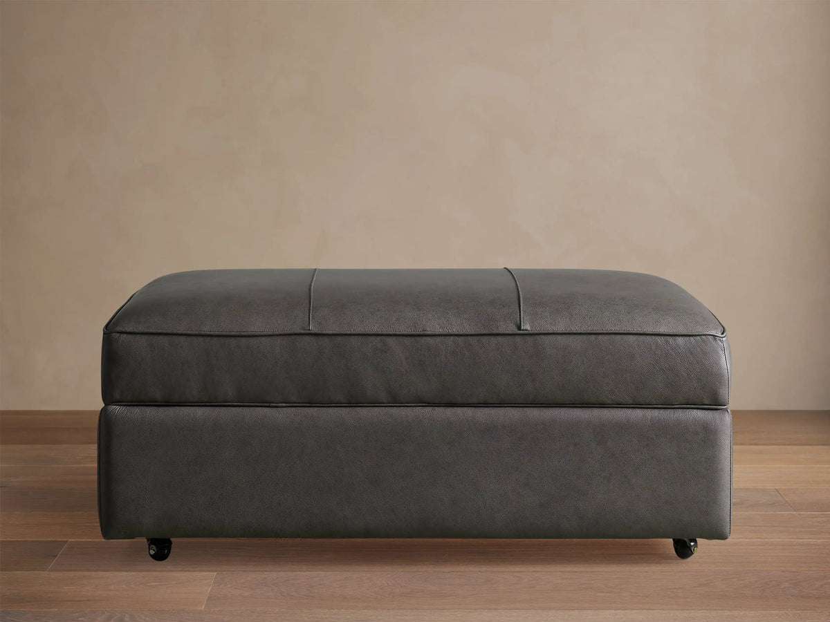 Filmore dark gray leather storage ottoman, rectangular, padded top with seams, on casters, against a neutral wall.