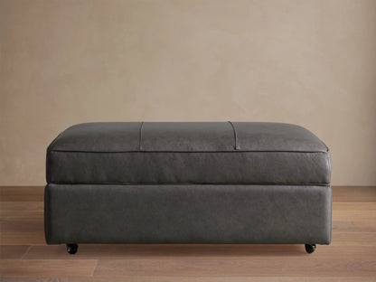 Filmore dark gray leather storage ottoman, rectangular, padded top with seams, on casters, against a neutral wall.