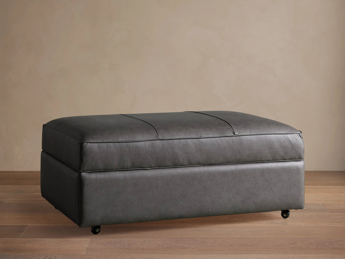 Filmore dark gray leather storage ottoman, rectangular, padded top with seams, on casters, against a neutral wall.