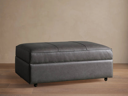 Filmore dark gray leather storage ottoman, rectangular, padded top with seams, on casters, against a neutral wall.
