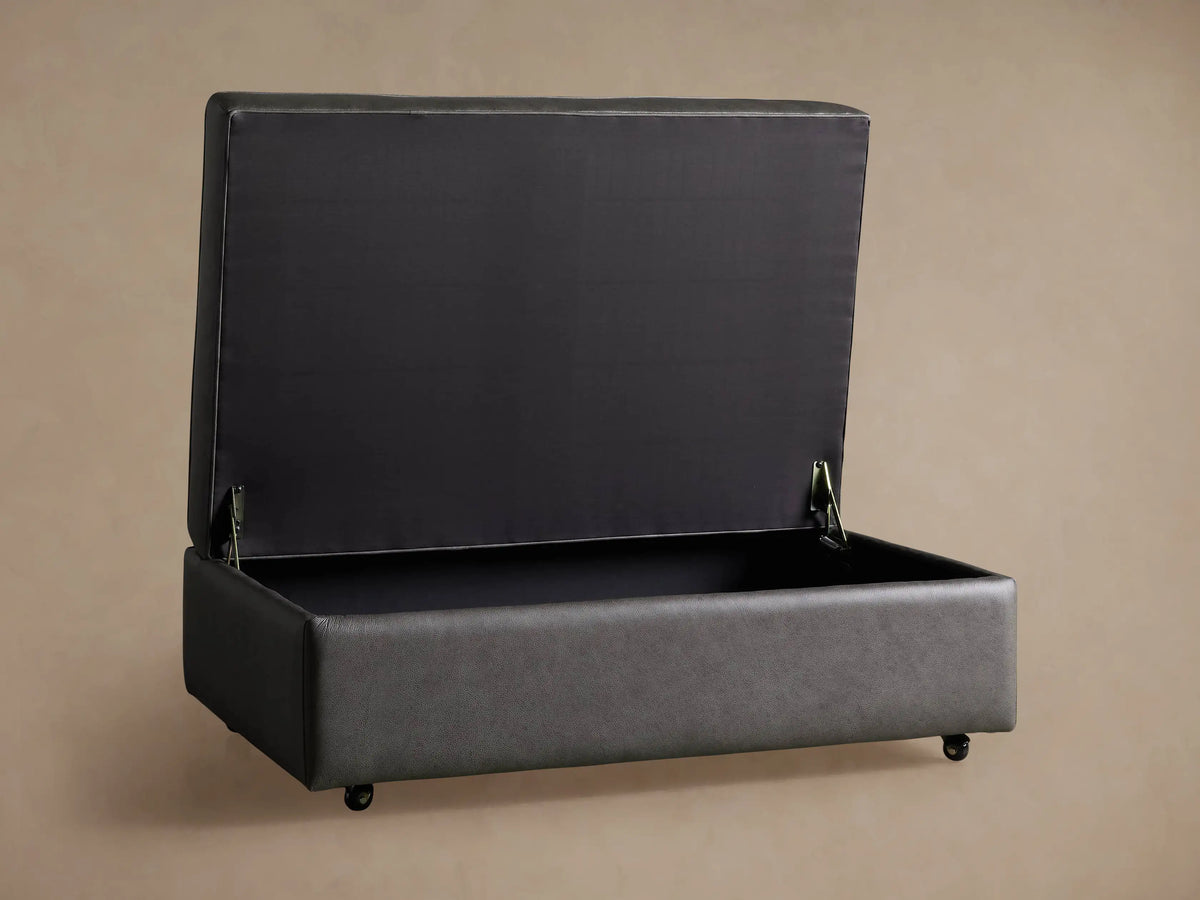 Close-up of open Filmore Leather Storage Ottoman. Angled view reveals dark interior lining & textured gray-brown leather exterior with hinge.