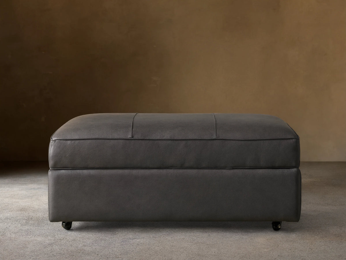 Filmore dark gray leather storage ottoman, rectangular, padded top with seams, on casters, against a neutral wall.