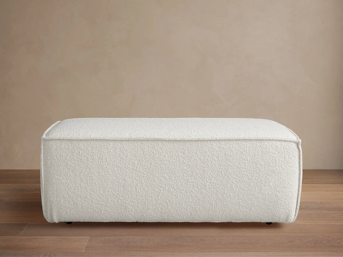 Front view of a rectangular Gabe Ottoman, upholstered in textured off-white fabric, on a wooden floor against a beige wall.