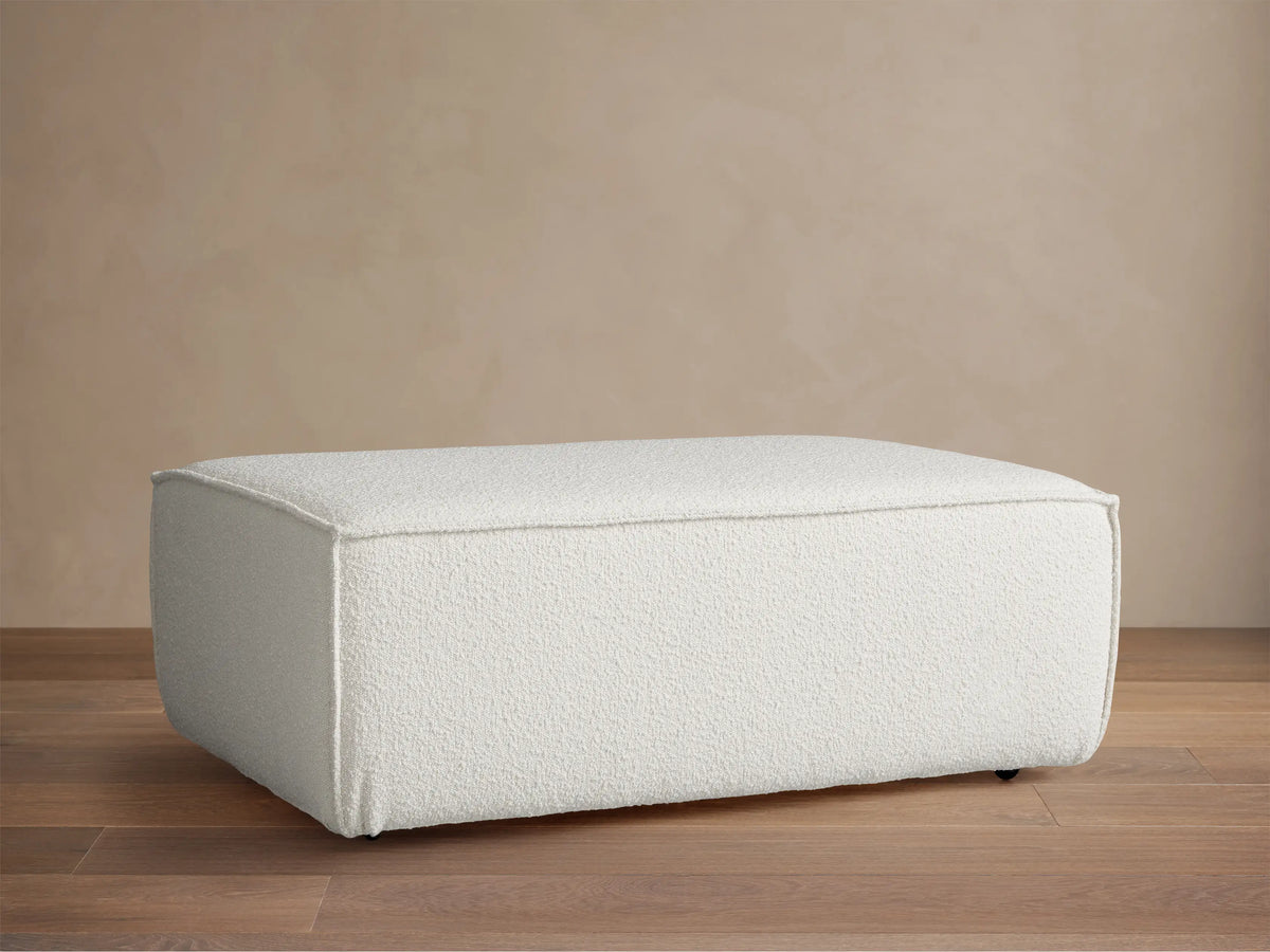 Front view of a rectangular Gabe Ottoman, upholstered in textured off-white fabric, on a wooden floor against a beige wall.