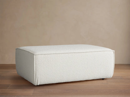 Front view of a rectangular Gabe Ottoman, upholstered in textured off-white fabric, on a wooden floor against a beige wall.