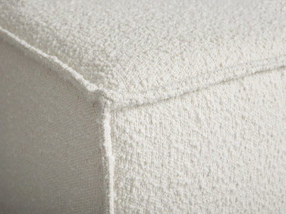 Rectangular Gabe Ottoman in cream bouclv© fabric with piping, on a wooden floor against a light beige wall.