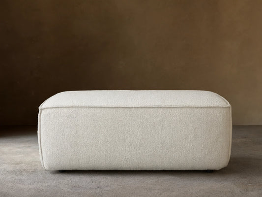 Front view of a rectangular Gabe Ottoman, upholstered in textured off-white fabric, on a wooden floor against a beige wall.