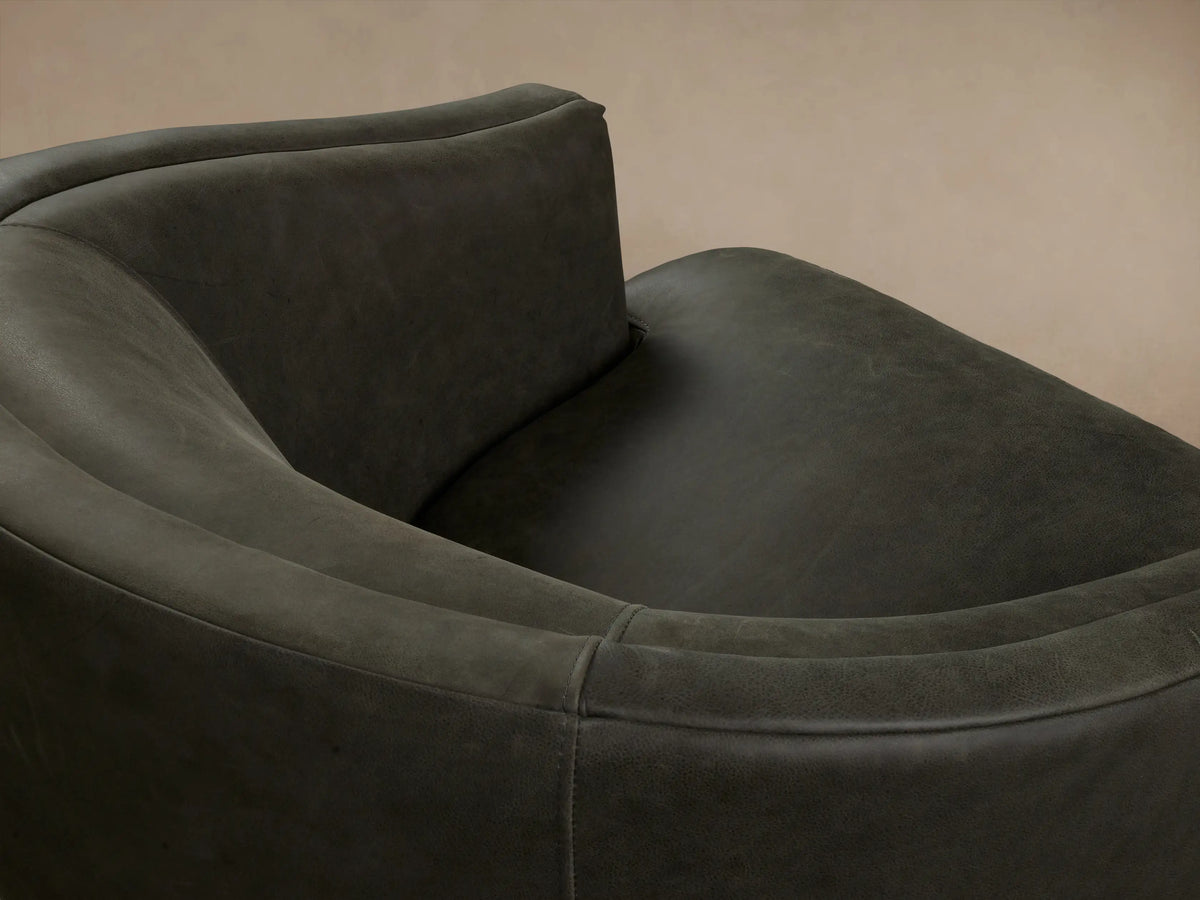 Dark green Kieran Leather Swivel Chair with a rounded backrest and cushioned seat, shown on wooden floor against a beige wall.