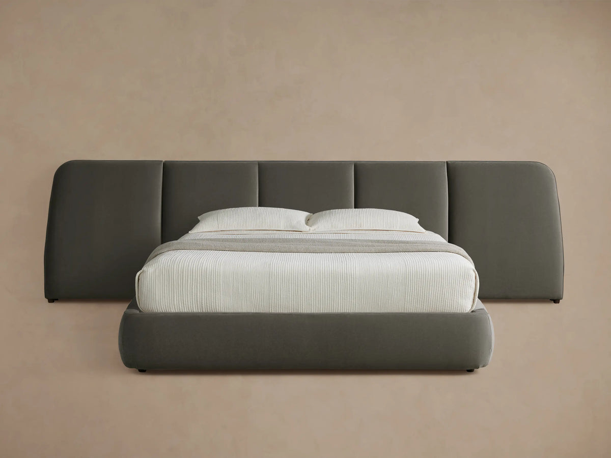 A modern bed with a wide padded headboard in dark gray fabric. It has a white mattress, two pillows, and a gray throw blanket on a wood floor.
