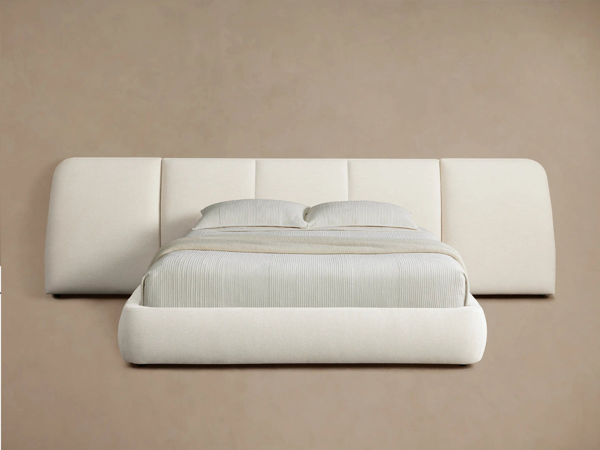 A modern cream-colored bed with a low profile and wide cushioned headboard, set against a beige wall, on wooden flooring.