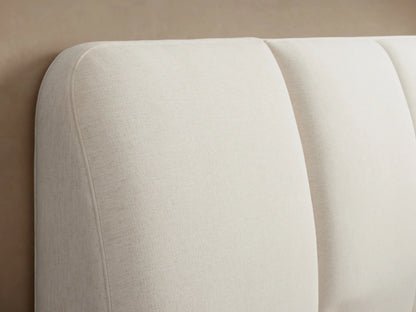A modern, beige upholstered bed with a wide, padded headboard and neatly made bedding against a neutral wall, viewed from the front.