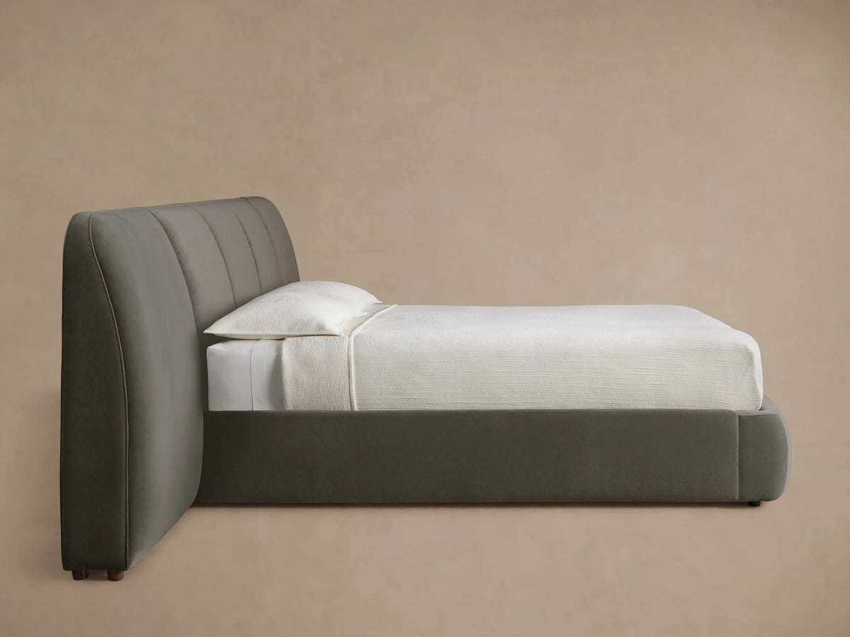 A close-up of a modern, dark gray upholstered headboard with smooth, rounded vertical panels against a light beige wall.