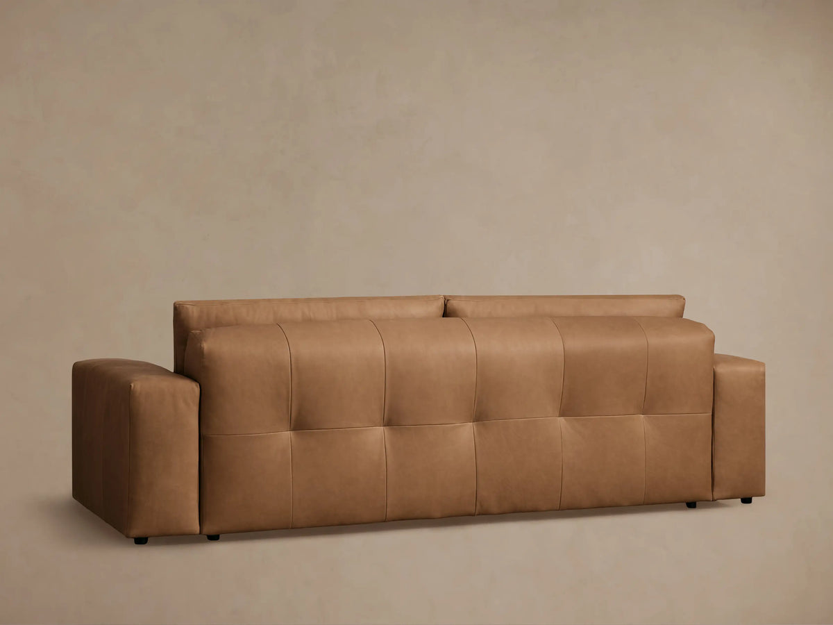 Close-up of a warm brown Keegan Leather Sofa, showing smooth, tufted seat cushions with stitching and a visible armrest.