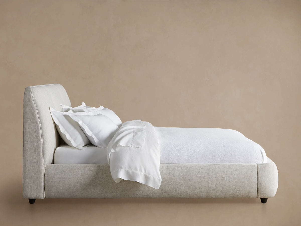 Kinnan Bed featuring an upholstered, cushioned headboard and a sleek, modern frame in a soft beige fabric. It rests on a wooden floor.