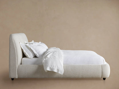 Kinnan Bed featuring an upholstered, cushioned headboard and a sleek, modern frame in a soft beige fabric. It rests on a wooden floor.