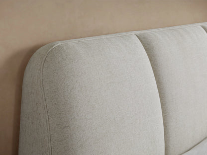 Kinnan Bed in beige fabric, side view. Features a tall, angled headboard with crisp white bedding tucked neatly, on dark wood legs.