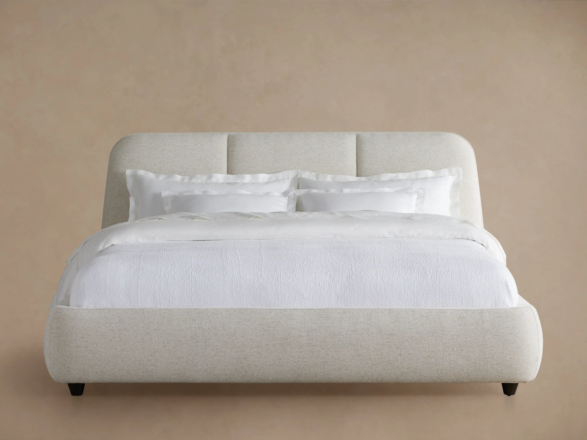 Cream-colored upholstered headboard of the Kinnan Bed, featuring soft fabric with subtle texture and rounded edges against a neutral wall.