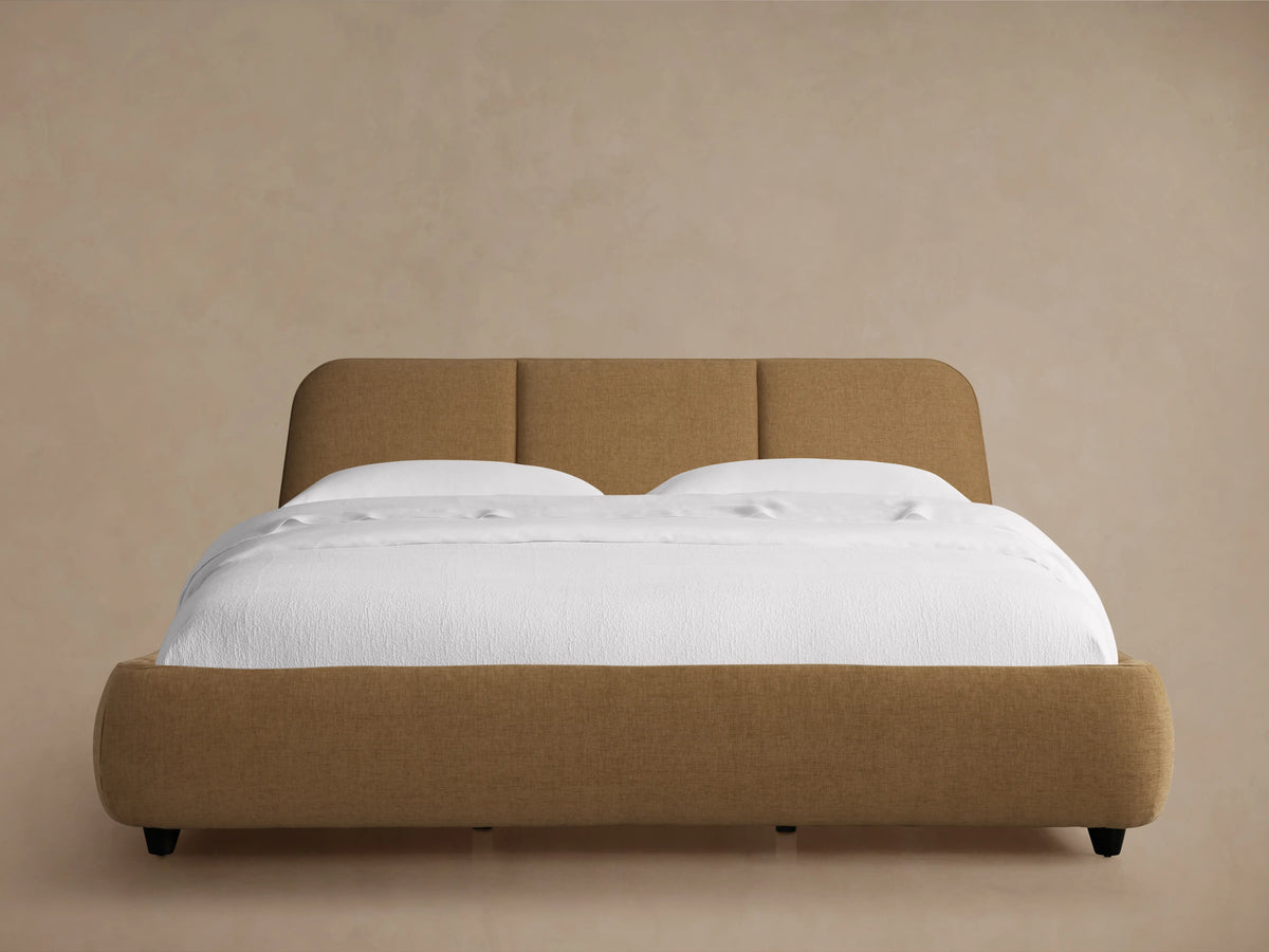Kinnan Bed in tan fabric with a padded headboard, set against a neutral wall. White bedding is neatly arranged on the mattress.