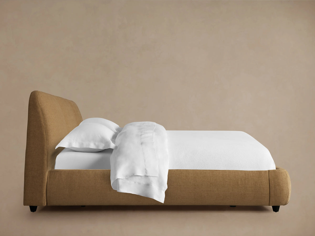 Kinnan Bed in a warm brown upholstery, features a three-panel headboard and rounded corners, shown with white bedding.