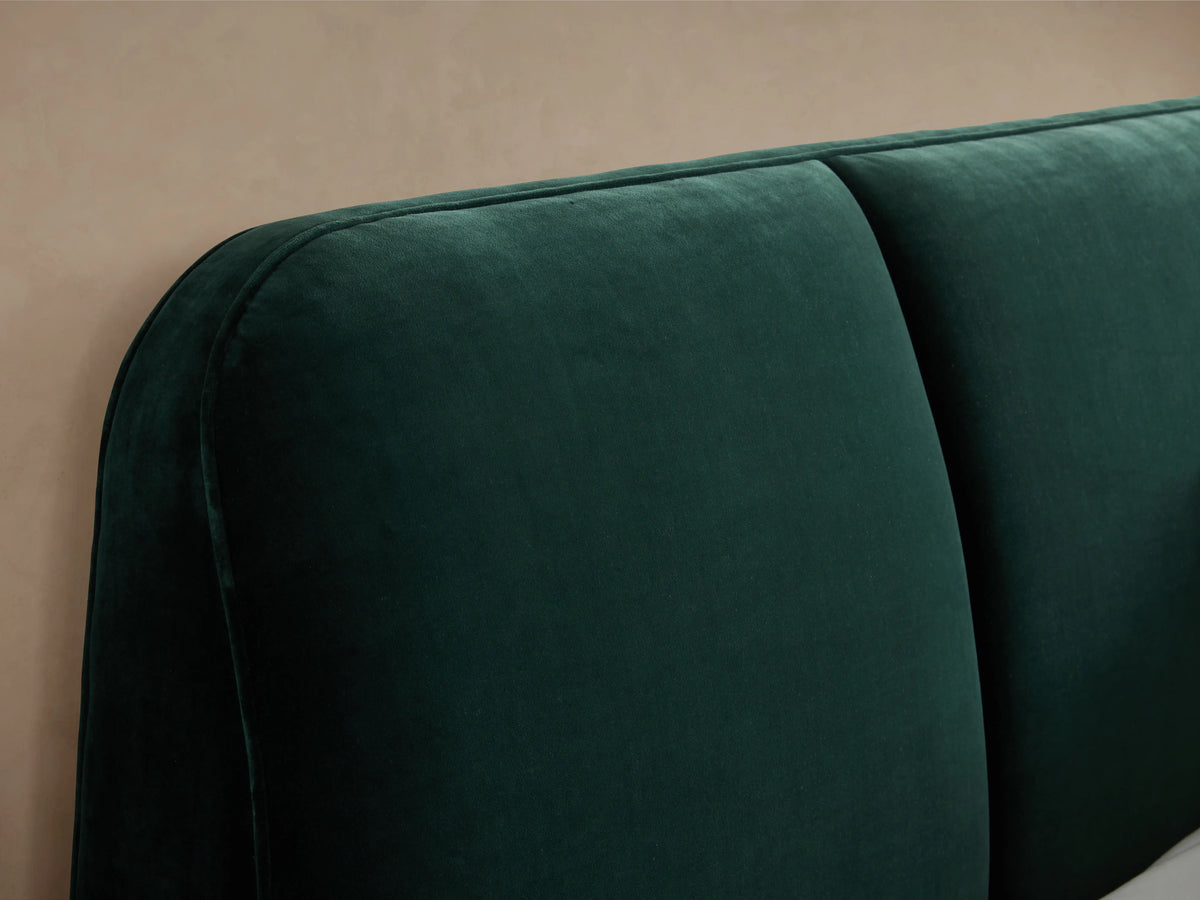 Dark green upholstered headboard of the Kinnan Bed with smooth, plush texture. The side view shows its rounded edges against a neutral wall background.