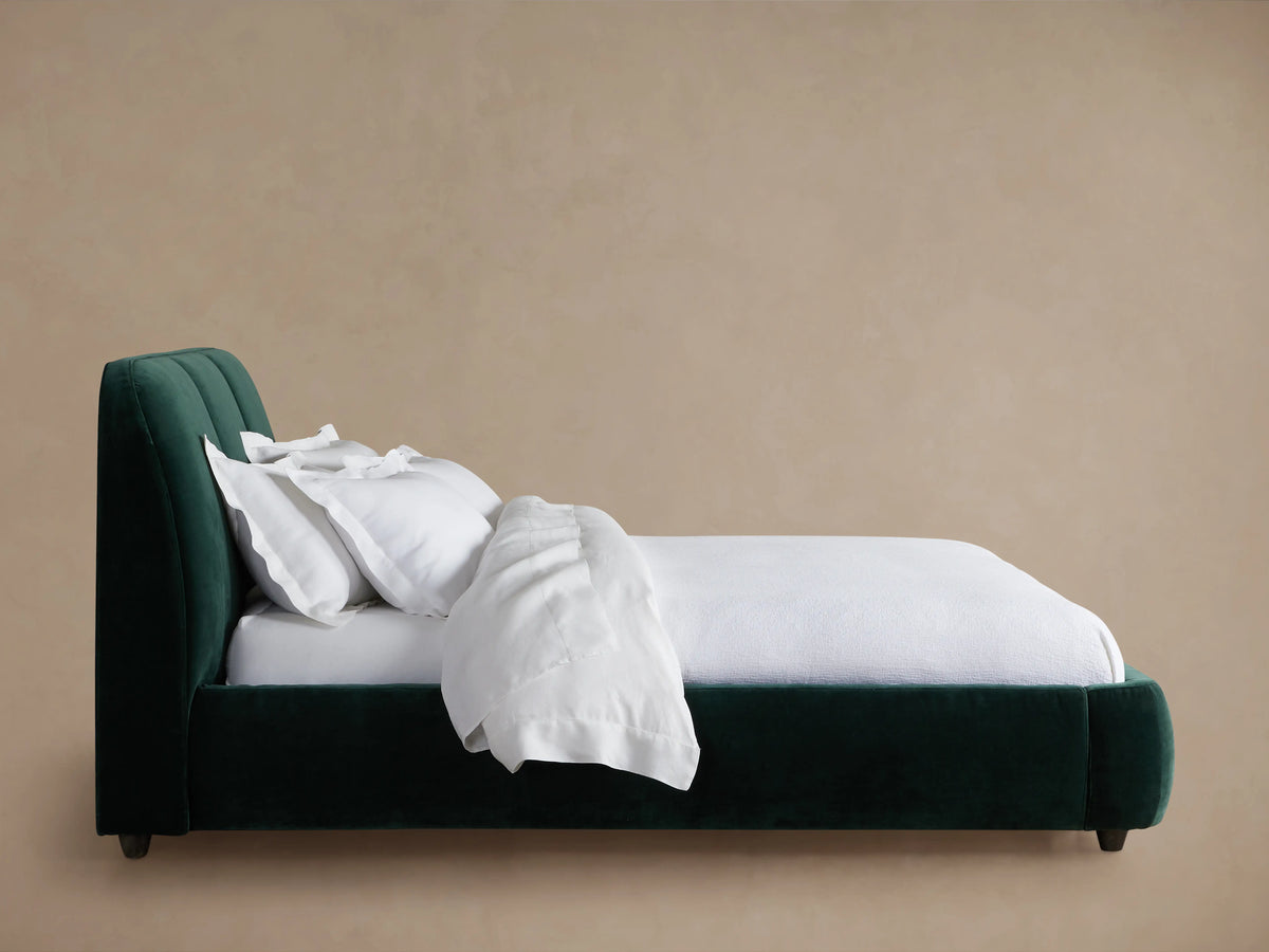 A plush Kinnan Bed with a dark green upholstered frame, a tall, padded headboard, and white linens neatly arranged, set against a beige background.