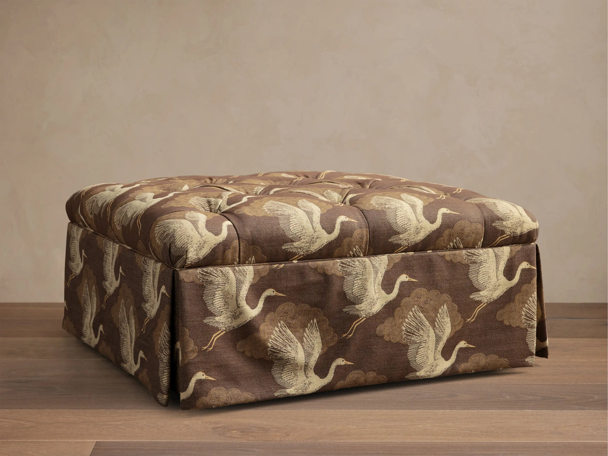 Front view of the brown Liddie Square Ottoman with a goldish-beige crane and cloud pattern, featuring a tufted top and pleated skirt.