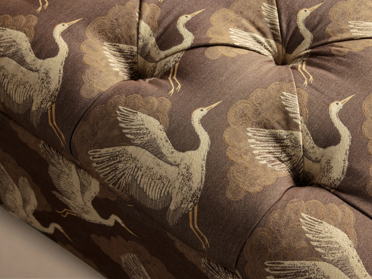 Angled view of a brown Liddie Square Ottoman, tufted top and pleated skirt, with off-white crane and brown cloud pattern.