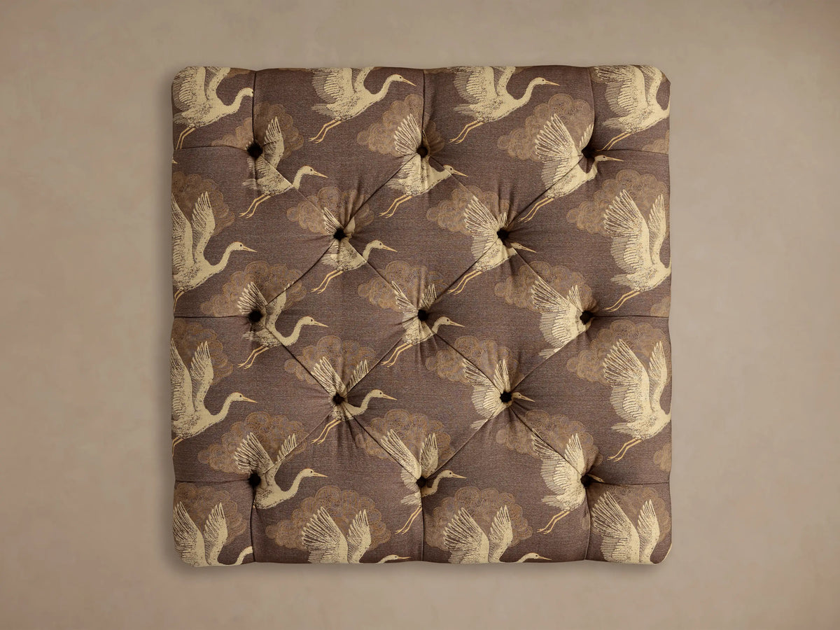 Close-up of the plum-brown Liddie Square Ottoman, showing its patterned upholstery of cream cranes and golden clouds, with tufting.