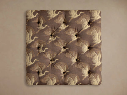 Close-up of the plum-brown Liddie Square Ottoman, showing its patterned upholstery of cream cranes and golden clouds, with tufting.