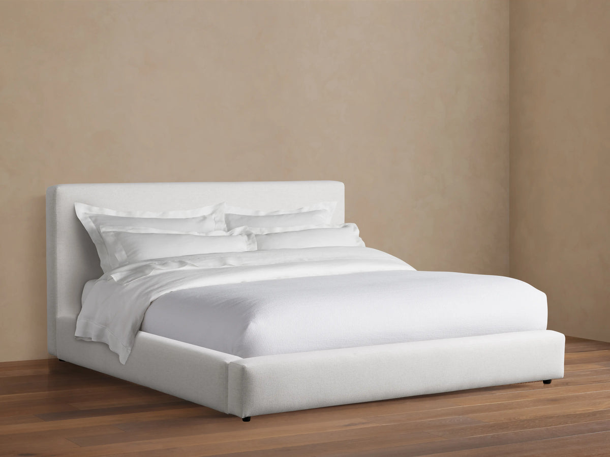 The Lago Bed features a simple, modern design with a white upholstered headboard and frame, set on a wooden floor against a beige wall.