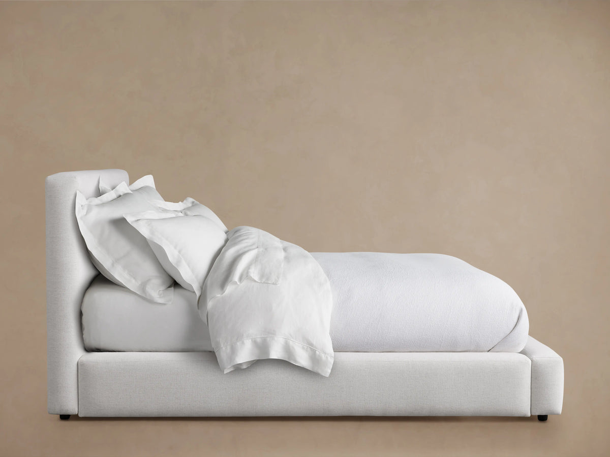 Lago Bed with light fabric upholstery, featuring a tall cushioned headboard. White bedding is slightly tousled on the mattress.