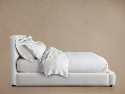 Lago Bed with light fabric upholstery, featuring a tall cushioned headboard. White bedding is slightly tousled on the mattress.