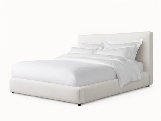 The Lago Bed features a simple, modern design with a white upholstered headboard and frame, set on a wooden floor against a beige wall.