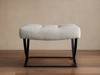 Light grey upholstered Lloyd Ottoman with two tufts and a black metal Z-shaped base, on a wooden floor.
