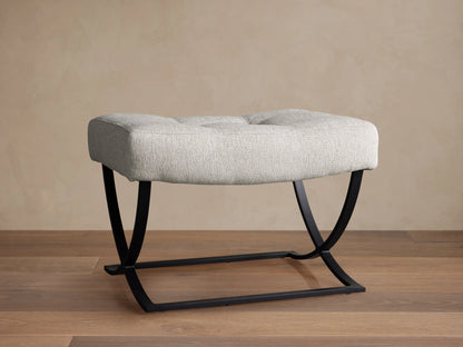 Light grey upholstered Lloyd Ottoman with two tufts and a black metal Z-shaped base, on a wooden floor.