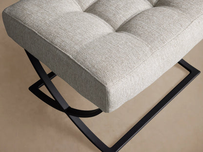 Lloyd Ottoman, light gray upholstered and tufted, featuring a black X-shaped metal base on a wooden floor.
