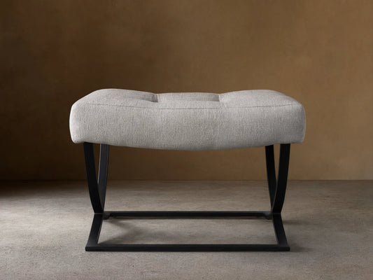 Light grey upholstered Lloyd Ottoman with two tufts and a black metal Z-shaped base, on a wooden floor.