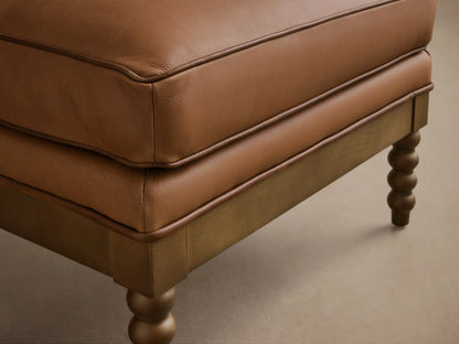 Front view of the Brown Milly Leather Ottoman with light wood spindled legs and visible cushion piping.